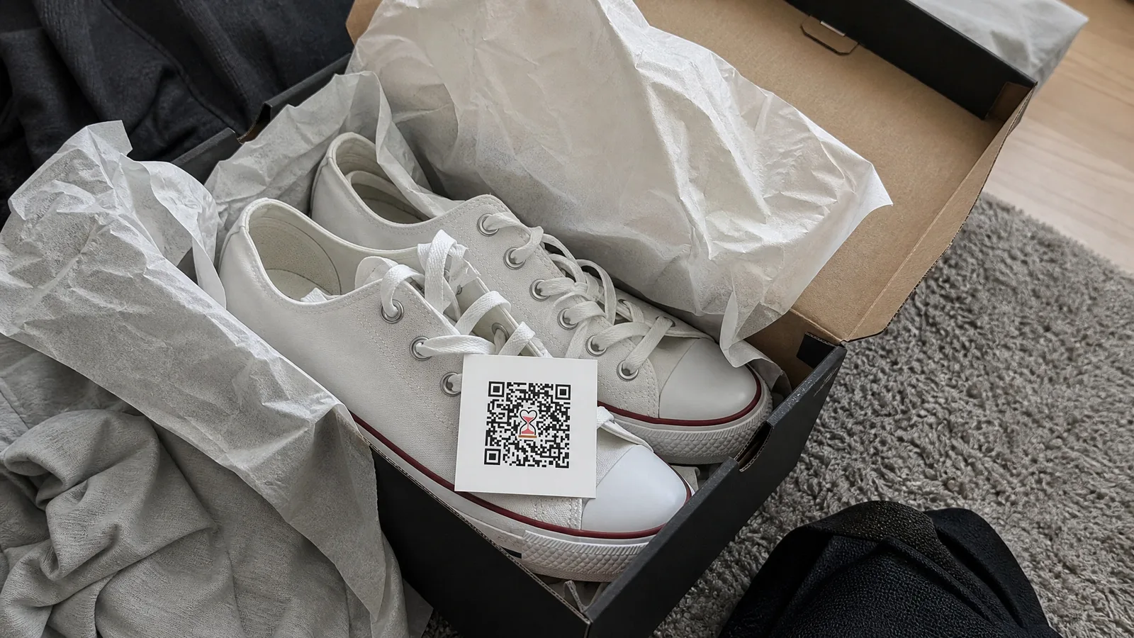 Caixa de tênis aberta com tênis casual branco, papel de seda amassado e cartão de QR Code apoiado sobre os tênis