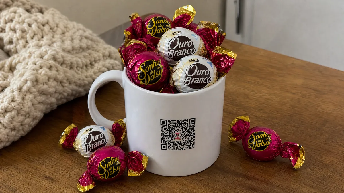 Caneca branca com QR Code estampado, cheia de bombons sortidos transbordando, alguns caídos ao redor