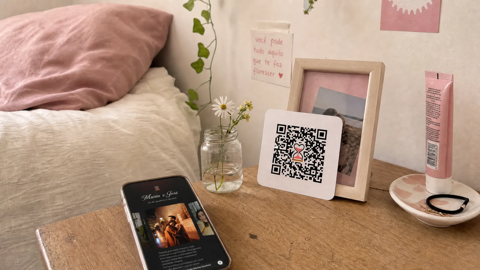 Cantinho de penteadeira simples com cartão de QR Code apoiado, flores do campo num potinho de vidro e celular mostrando a página do casal com cronômetro